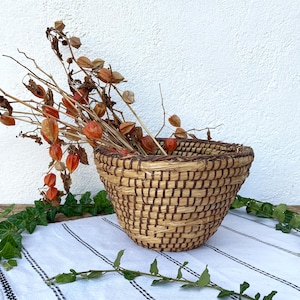 May include: A woven straw basket with a brown and tan pattern. The basket is filled with dried orange flowers and is sitting on a white and black striped tablecloth.