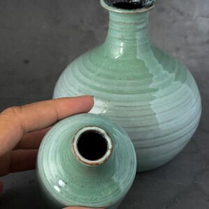 May include: Two ceramic vases in a light green glaze with a darker green rim. The larger vase has a round body and a narrow neck. The smaller vase is round with a slightly wider neck.