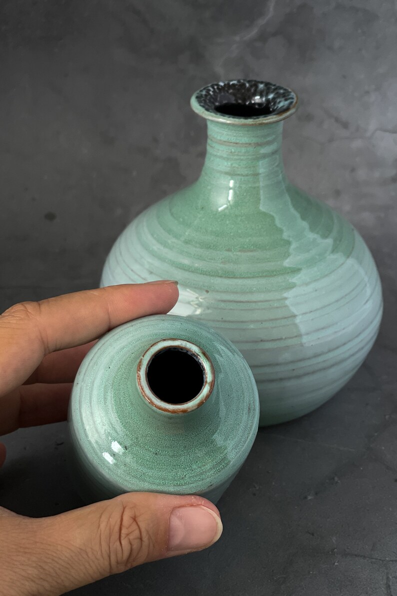 May include: Two ceramic vases in a light green glaze with a darker green rim. The larger vase has a round body and a narrow neck. The smaller vase is round with a slightly wider neck.