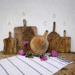 Puede incluir: Cinco tablas de cortar de madera antiguas de varias formas y tamaños están dispuestas sobre una superficie de madera. Las tablas están desgastadas con vetas de madera visibles e imperfecciones naturales. Un paño a rayas blancas y negras está en primer plano, con flores rosas.