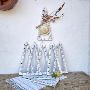 以下が含まれることがあります： クリーム色の蓋とドライフラワーアレンジメントが付いた、白く風化した金属製の壁掛け式鍋ラック。木製のテーブルに、模様入りの布と小さなカボチャと一緒に、お揃いのラックがいくつか配置されています。素朴なキッチンインテリア。