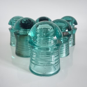 Pode incluir: Uma coleção de seis isoladores elétricos de vidro vintage em tom azul-petróleo dispostos em grupo. Cada isolador apresenta um topo arredondado em forma de cúpula, uma crista central larga e uma base cilíndrica com detalhes canelados.
