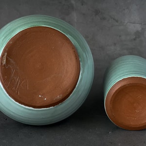 May include: Two ceramic pots with a light green glaze. The pots are unglazed on the bottom, revealing the brown clay. The larger pot has a signature on the bottom.