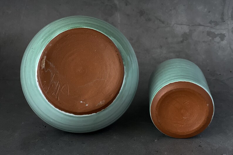May include: Two ceramic pots with a light green glaze. The pots are unglazed on the bottom, revealing the brown clay. The larger pot has a signature on the bottom.