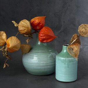 May include: Two teal ceramic vases with dried orange and brown flowers. The larger vase is wider and rounder than the smaller vase.