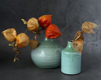 Op de afbeelding: Twee teal keramische vazen met gedroogde oranje en bruine bloemen. De grotere vaas is breder en ronder dan de kleinere vaas.