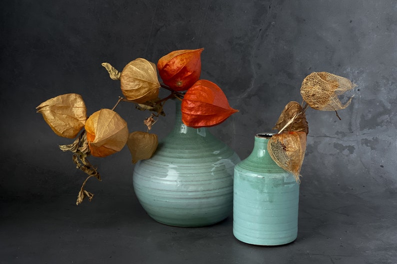 May include: Two teal ceramic vases with dried orange and brown flowers. The larger vase is wider and rounder than the smaller vase.