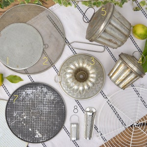 Op de afbeelding: Negen vintage metalen bakvormen en gereedschap, waaronder een ronde taartvorm, een tulbandvorm, twee kleine geribbelde vormen, een rond koelrek, een ronde pizzaplaat, een ronde draadgaasvorm en een rasp. De vormen zijn allemaal zilverkleurig en hebben een versleten, rustieke uitstraling.
