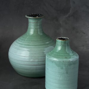 May include: Two ceramic vases in shades of green and blue. The larger vase has a round body and a narrow neck. The smaller vase is cylindrical. Both vases have a textured, glazed finish.