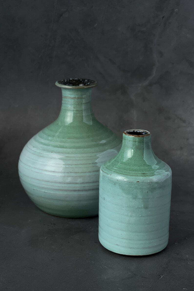 May include: Two ceramic vases in shades of green and blue. The larger vase has a round body and a narrow neck. The smaller vase is cylindrical. Both vases have a textured, glazed finish.