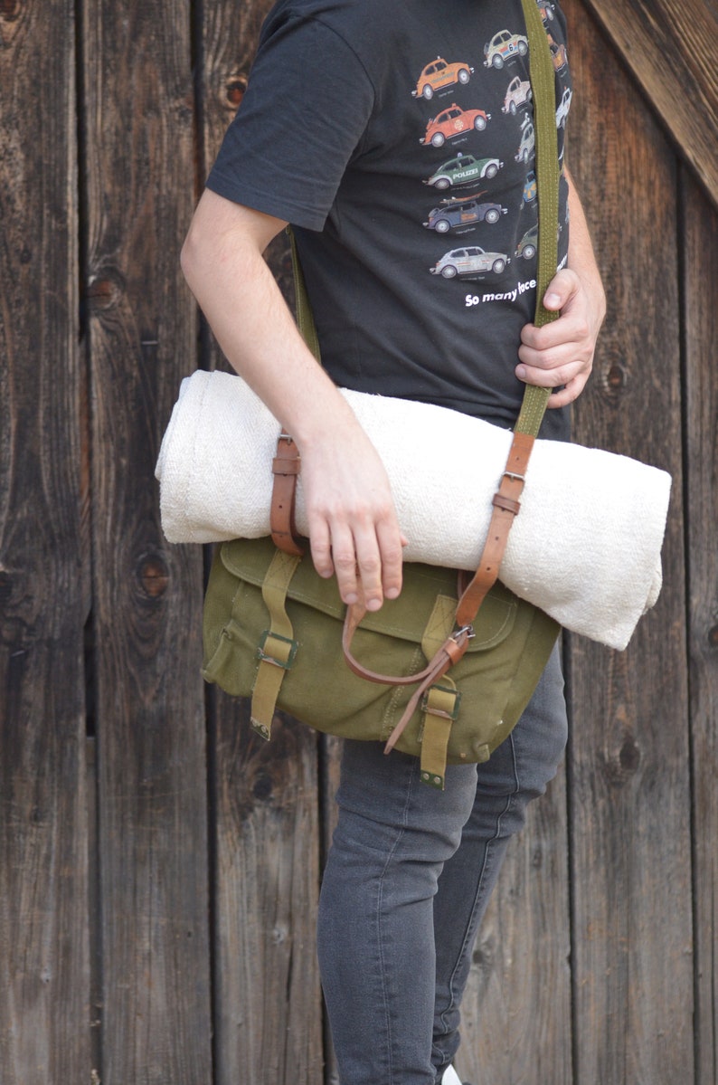May include: A person wearing a black t-shirt with a print of various Volkswagen Beetles and the text "So many ice". They are carrying a green canvas bag with a brown leather strap and a white towel.