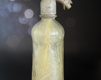 Pode incluir: Uma garrafa de seltzer de vidro antigo com um dispensador de metal. A garrafa transparente tem um tom amarelado e letras em relevo. O dispensador tem uma alavanca e um bico. O design é clássico e vintage.