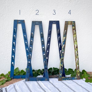 Puede incluir: Cuatro marcos de metal azul con ganchos, cada uno con un diseño diferente. Los marcos están dispuestos en una fila sobre una superficie blanca. Los números 1, 2, 3 y 4 están escritos encima de cada marco.