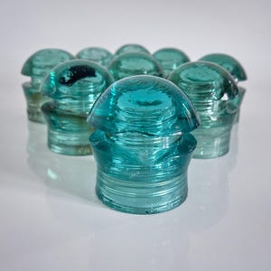 Puede incluir: Una colección de diez aisladores eléctricos de vidrio vintage en color verde azulado. Cada pieza presenta una parte superior en forma de cúpula redondeada, una ranura central y una base cilíndrica. El vidrio es translúcido y pulido.