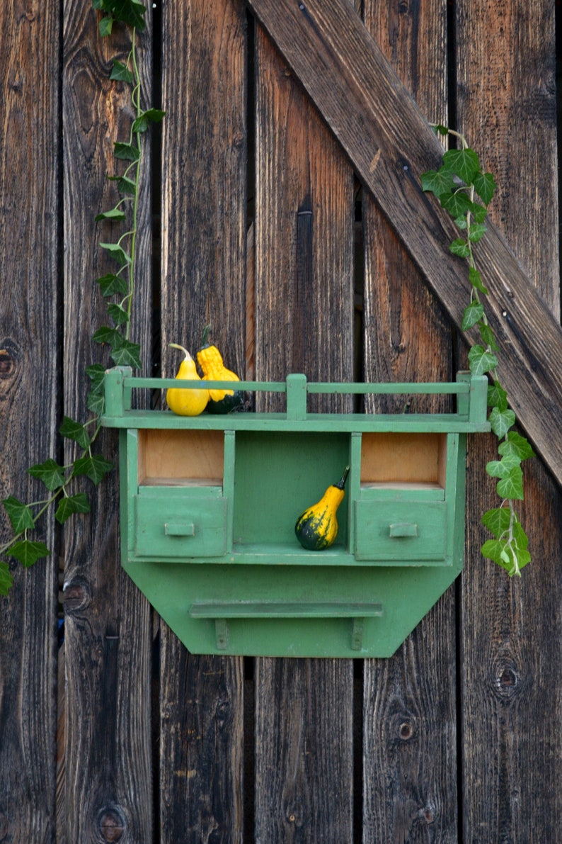 May include: A green wooden shelf with three drawers and a small shelf on the bottom. The shelf is mounted on a wooden wall and has three gourds on the top shelf.