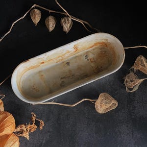 Op de afbeelding: Een ovale, witte metalen bakvorm met roestkleurige vlekken. De lege vorm staat op een donkere ondergrond, met gedroogde plantenelementen. De bakvorm heeft een vintage, rustieke uitstraling.
