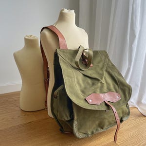 May include: An olive green canvas backpack with brown leather accents. The backpack has a flap closure, a front pocket, and adjustable leather straps. The bag is displayed on a mannequin.
