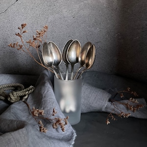 Puede incluir: Un grupo de cucharas de plata en un jarrón de vidrio esmerilado sobre una superficie de tela gris. Las cucharas están dispuestas en forma de abanico con los mangos apuntando hacia arriba. El jarrón está sobre una superficie de tela gris.