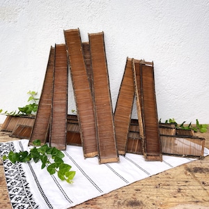 Puede incluir: Varios bandejas de bambú tejido marrón, de forma rectangular y ligeramente inclinadas, se exhiben. Se disponen sobre un paño blanco con un borde con estampado en blanco y negro, adornadas con follaje verde.