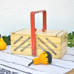 May include: A vintage wooden sewing box with a red handle and metal accents. The box is closed and has a white finish. Two small gourds are in the foreground.