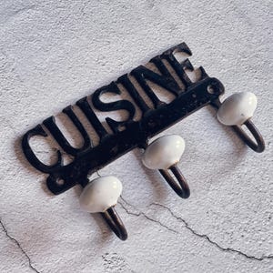 Könnte beinhalten: Ein schwarzes Metallschild mit dem Wort "Cuisine" in weißen Buchstaben. Das Schild hat drei weiße Keramikhaken.
