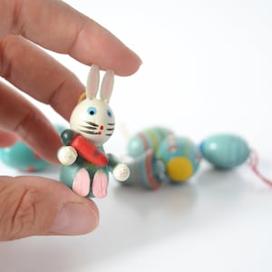 May include: A white wooden bunny rabbit ornament with blue and pink painted details holding a red carrot. The bunny is part of a string of Easter egg ornaments, with blue and yellow painted details.