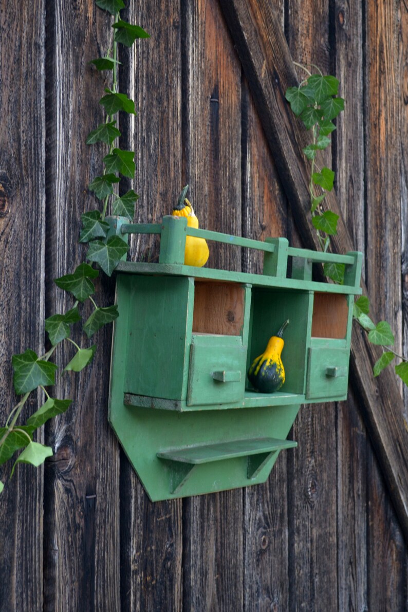May include: A green wooden shelf with three compartments and a lower shelf. The shelf is attached to a wooden wall and has two gourds on it, one yellow and one green.