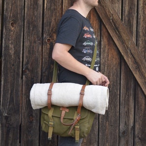 May include: A person wearing a black t-shirt with a vintage car print and a green canvas bag with brown leather straps. The bag is slung over the person's shoulder and has a white blanket rolled up inside.