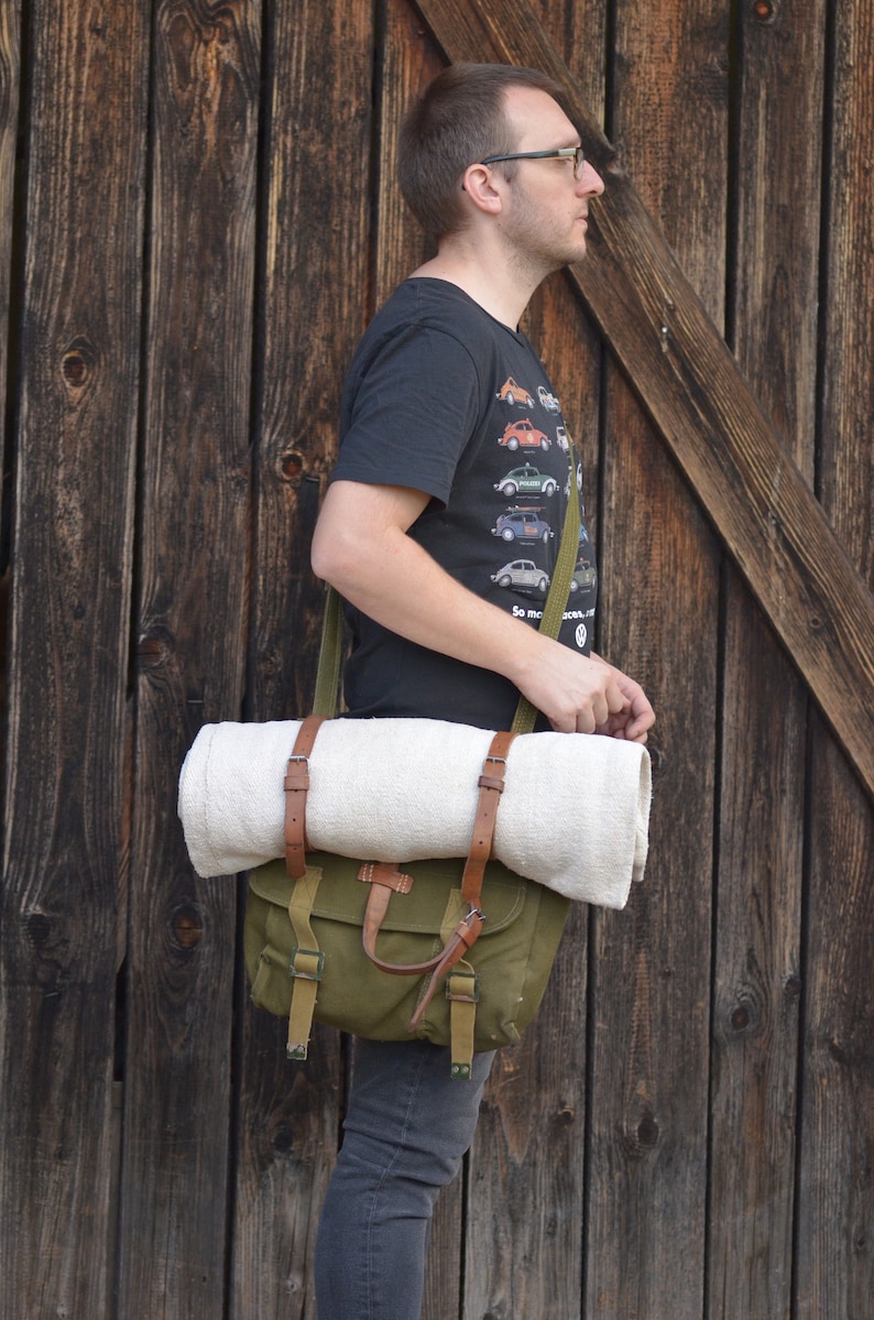 May include: A person wearing a black t-shirt with a vintage car print and a green canvas bag with brown leather straps. The bag is slung over the person's shoulder and has a white blanket rolled up inside.