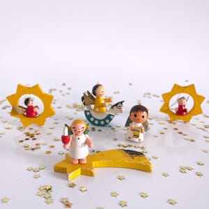 Könnte beinhalten: Eine Sammlung von Vintage-Weihnachtsschmuck mit kleinen Holzengelfiguren und sternförmigen Dekorationen. Die Engel sind in Weiß, Rot und Gelb gekleidet, mit goldenen Flügeln und halten Kerzen. Die Ornamente sind auf einer weißen Oberfläche mit goldenen Sternenkonfetti verstreut.