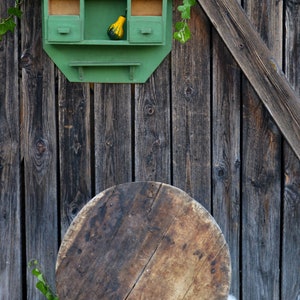 May include: A green wooden shelf with two drawers and a small ledge, mounted on a weathered wooden wall. The shelf is decorated with small gourds. A large, round wooden disc sits on a white fluffy surface in front of the shelf.
