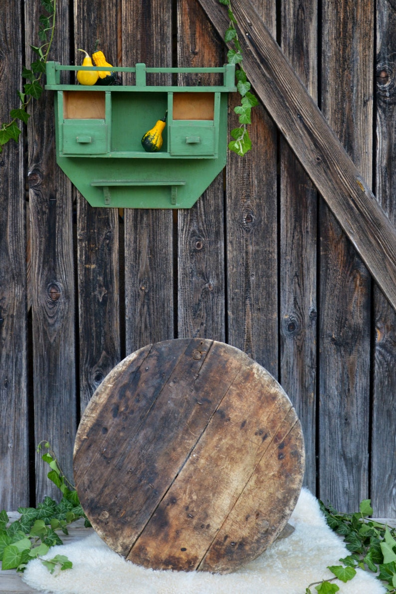 May include: A green wooden shelf with two drawers and a small ledge, mounted on a weathered wooden wall. The shelf is decorated with small gourds. A large, round wooden disc sits on a white fluffy surface in front of the shelf.