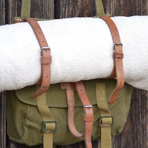 May include: A green canvas bag with brown leather straps and buckles. The bag is hanging on a wooden wall and has a white blanket inside.