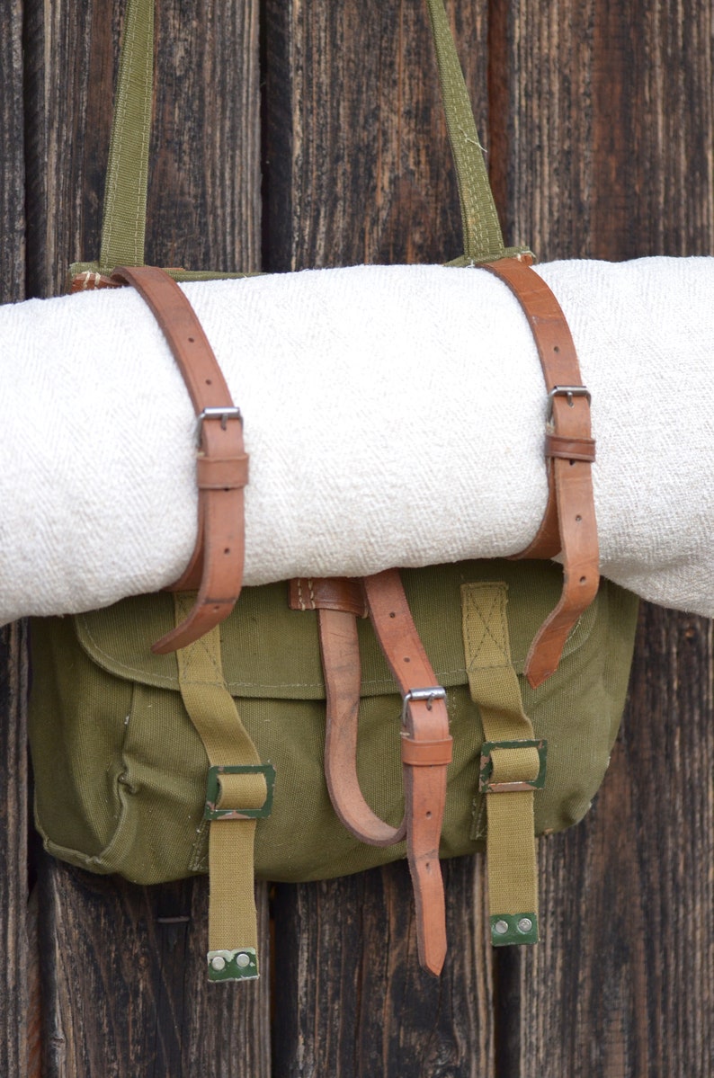 May include: A green canvas bag with brown leather straps and buckles. The bag is hanging on a wooden wall and has a white blanket inside.