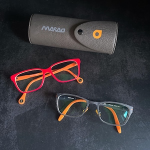 Puede incluir: Dos pares de gafas con monturas naranjas y rojas, y un estuche gris para gafas. Un par tiene monturas rojas con patillas naranjas, y el otro tiene una montura con estampado negro y gris con patillas naranjas. El estuche es gris con la marca "MASAO".