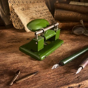 May include: A vintage green metal paper punch with the text "HERDEGEN 910" is displayed on a wooden surface. The image also shows a green pen, a pencil, and antique books and papers, suggesting a stationery or office theme.