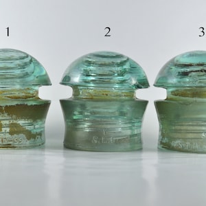 May include: Three vintage glass insulators in a light green colour. The insulators are numbered 1, 2, and 3. The insulators have a rounded top and a wide base. The base of each insulator has a different inscription. Insulator 1 says "J.T.S. 311-59", insulator 2 says "S.L.E.", and insulator 3 says "S.L.A. 59".