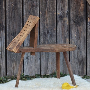 May include: A rustic wooden stool with three legs and a unique, angled top. The top is covered in small, evenly spaced holes. The stool is sitting on a white, fluffy rug in front of a wooden wall.