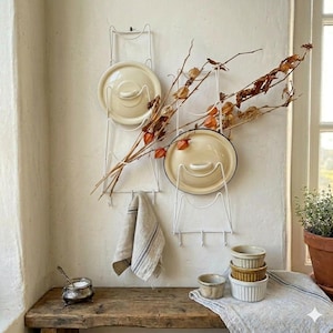 May include: A rustic kitchen scene features white wire pot lid racks holding cream-coloured lids. A sprig of dried orange-toned flowers and a linen towel hang nearby. A wooden bench holds small ceramic bowls and a silver salt cellar.
