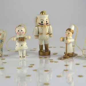 Könnte beinhalten: Fünf weiße Holz-Weihnachtsornamente mit goldenen Akzenten. Die Ornamente sind eine Glocke, ein Mädchen, das Akkordeon spielt, ein Nussknacker, ein Junge beim Skifahren und eine Glocke mit einem Stern. Die Ornamente befinden sich auf einer weißen Oberfläche mit goldenem Glitzer.