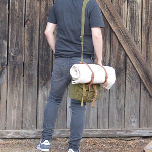 May include: A person wearing a black t-shirt and blue jeans is carrying a green canvas bag with a white rolled blanket inside. The bag has brown leather straps and is worn over the shoulder.