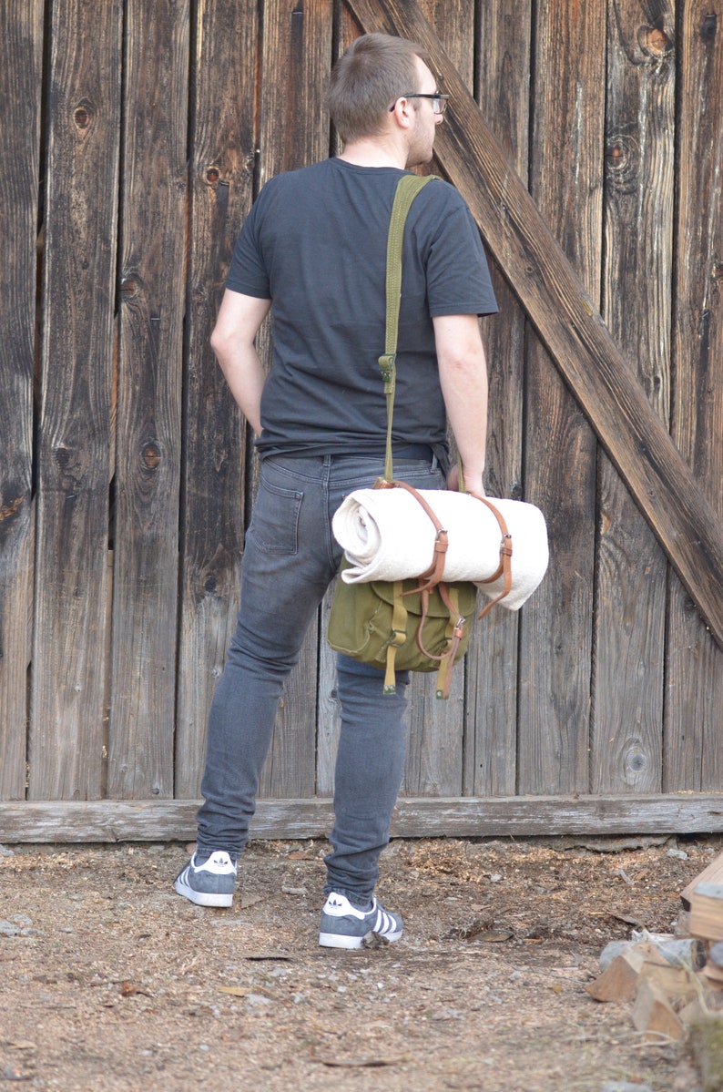 May include: A person wearing a black t-shirt and blue jeans is carrying a green canvas bag with a white rolled blanket inside. The bag has brown leather straps and is worn over the shoulder.