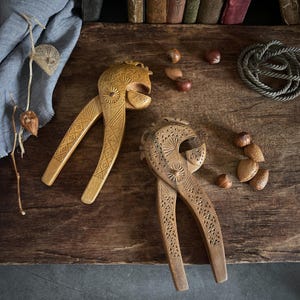 May include: Two light brown wooden nutcrackers featuring intricate geometric carvings and sunburst patterns on the handles and heads. The nutcrackers are positioned alongside several whole nuts, including almonds and hazelnuts, on a dark wood surface.