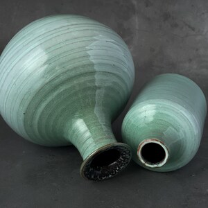May include: Two ceramic vases in a light green glaze. The larger vase has a rounded body and a narrow neck. The smaller vase is cylindrical. Both vases have a smooth, slightly textured surface.