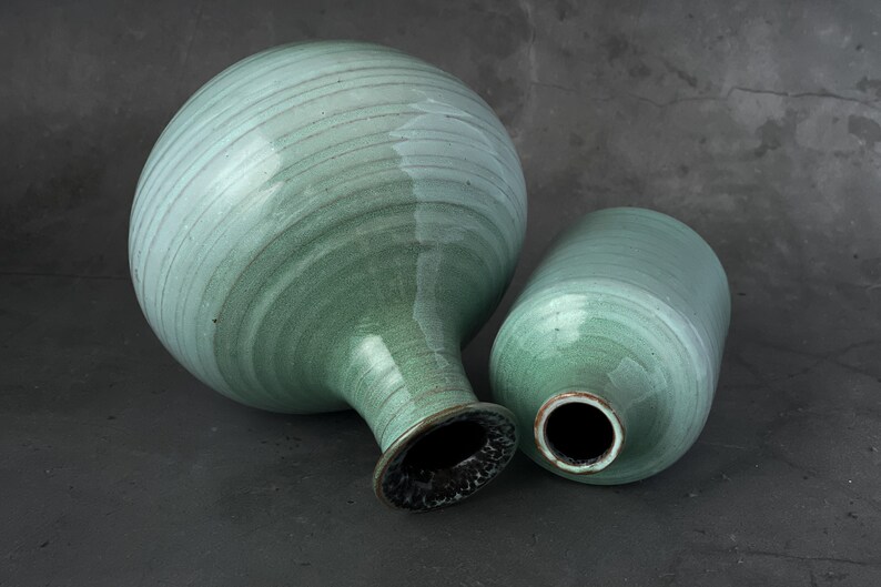 May include: Two ceramic vases in a light green glaze. The larger vase has a rounded body and a narrow neck. The smaller vase is cylindrical. Both vases have a smooth, slightly textured surface.