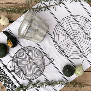 Op de afbeelding: Twee ronde draadkoelrekken, een metalen taartvorm en pompoenen op een wit en zwart gestreepte keukendoek.