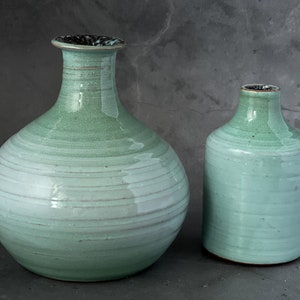 May include: Two ceramic vases in a light green glaze with a white stripe pattern. The larger vase has a round body and a narrow neck. The smaller vase is cylindrical.