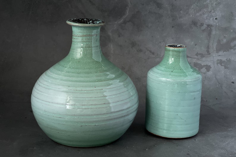 May include: Two ceramic vases in a light green glaze with a white stripe pattern. The larger vase has a round body and a narrow neck. The smaller vase is cylindrical.