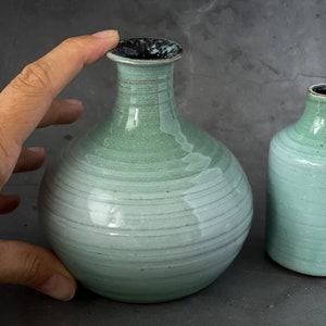 May include: Two ceramic vases in a light green glaze with a subtle white stripe pattern. The larger vase has a rounder body and a slightly wider opening. The smaller vase has a more cylindrical shape.