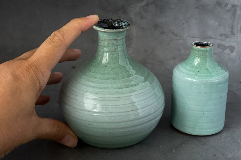 May include: Two ceramic vases in a light green glaze with a subtle white stripe pattern. The larger vase has a rounder body and a slightly wider opening. The smaller vase has a more cylindrical shape.